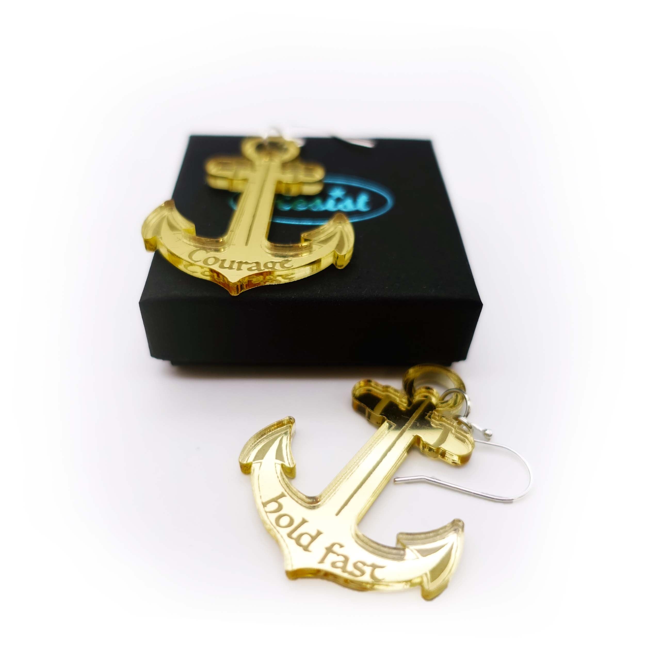 Pair of gold mirror gold anchor earrings etched with the words courage and hold fast on a Wear and Resist gift box in aid of the RNLI and Women for Refugee Women. 