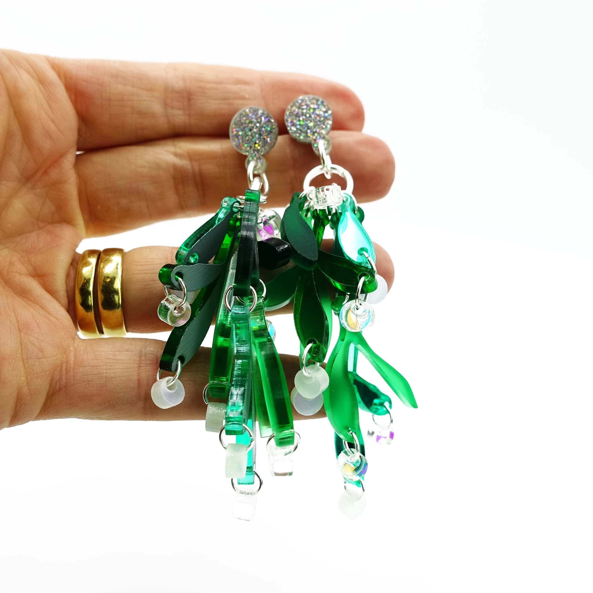 Hand holds mistletoe earrings with glitter tops and iridescent and opalescent white berries with mirror and frost green leaves. 