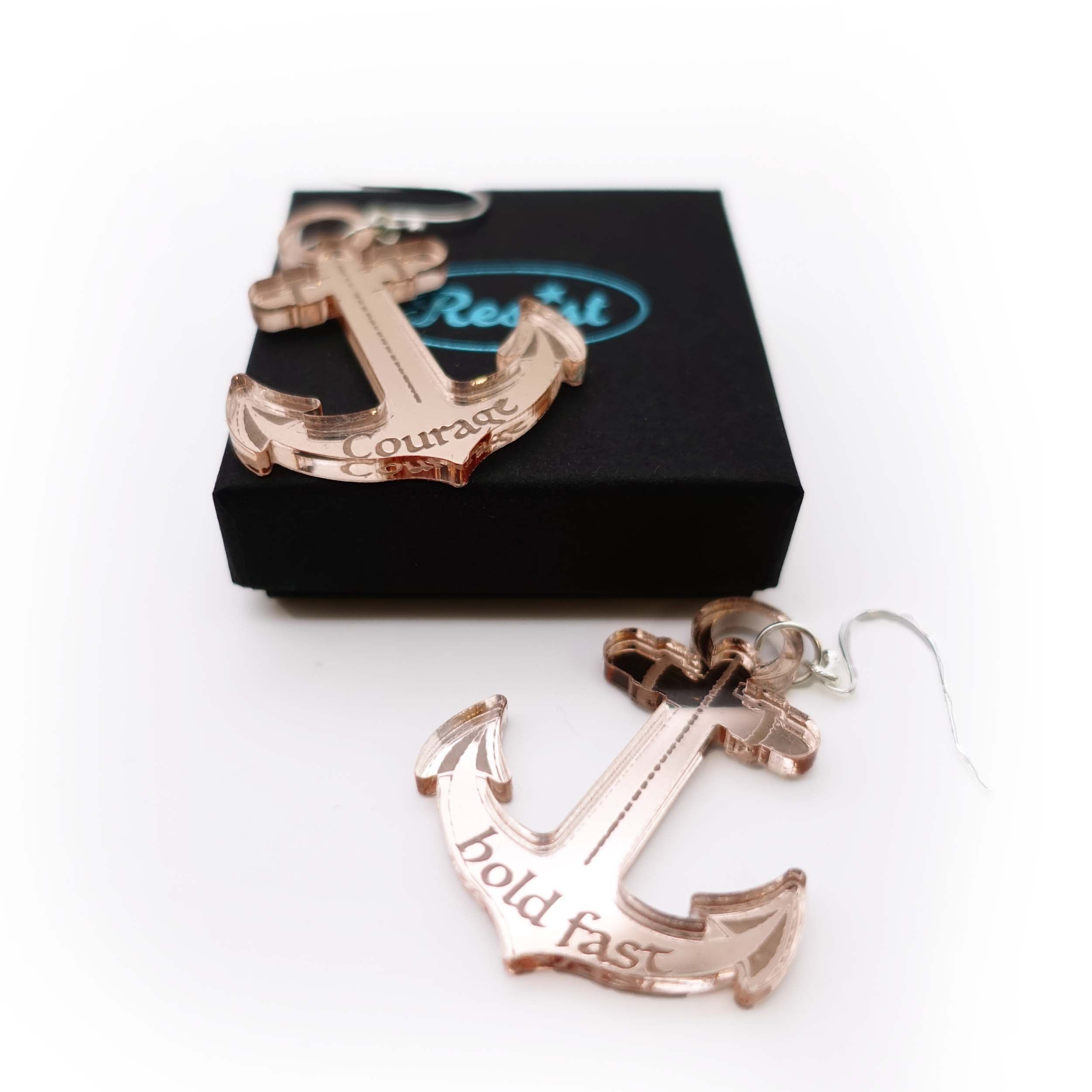 A pair of rose-gold mirror anchor earrings one etched with courage, the other etched with hold fast shown with a Wear and Resist gift box, sold with £2 going to the RNLI and another £2 going to Women for Refugee Women.
