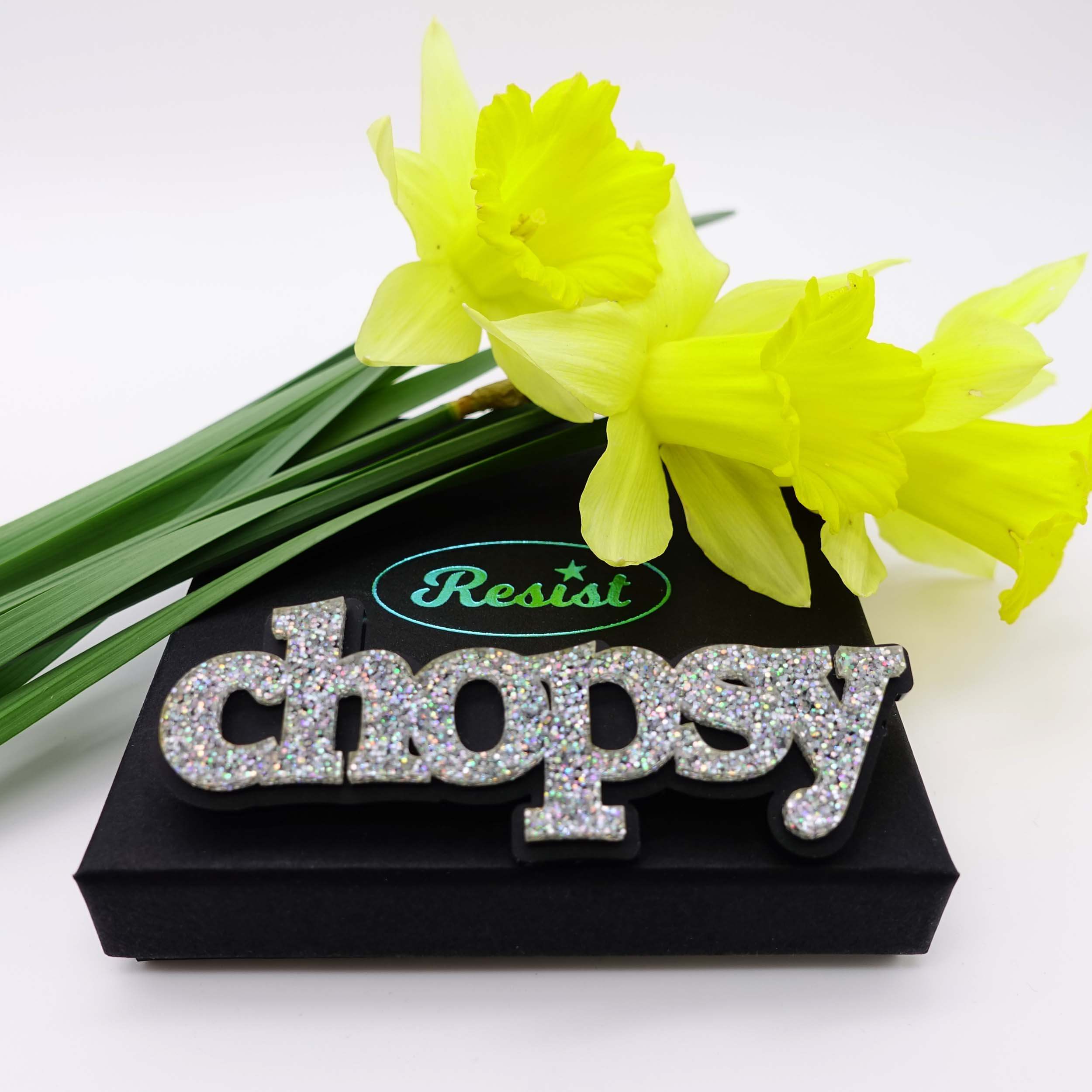 An iridescent silver glitter Chopsy brooch designed by Sarah Day for Wear and Resist, shown on a gift box with daffodils. £2 goes to Welsh Women's Aid. Fel yr arfer, mae £2 o bob mwclus Chopsy yn mynd at @welshwomensaid.