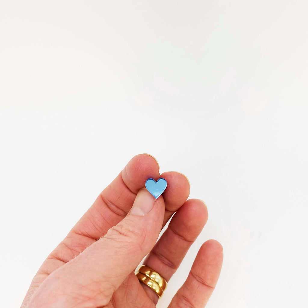 A hand shown holding a sky blue tiny heart stud earring. 