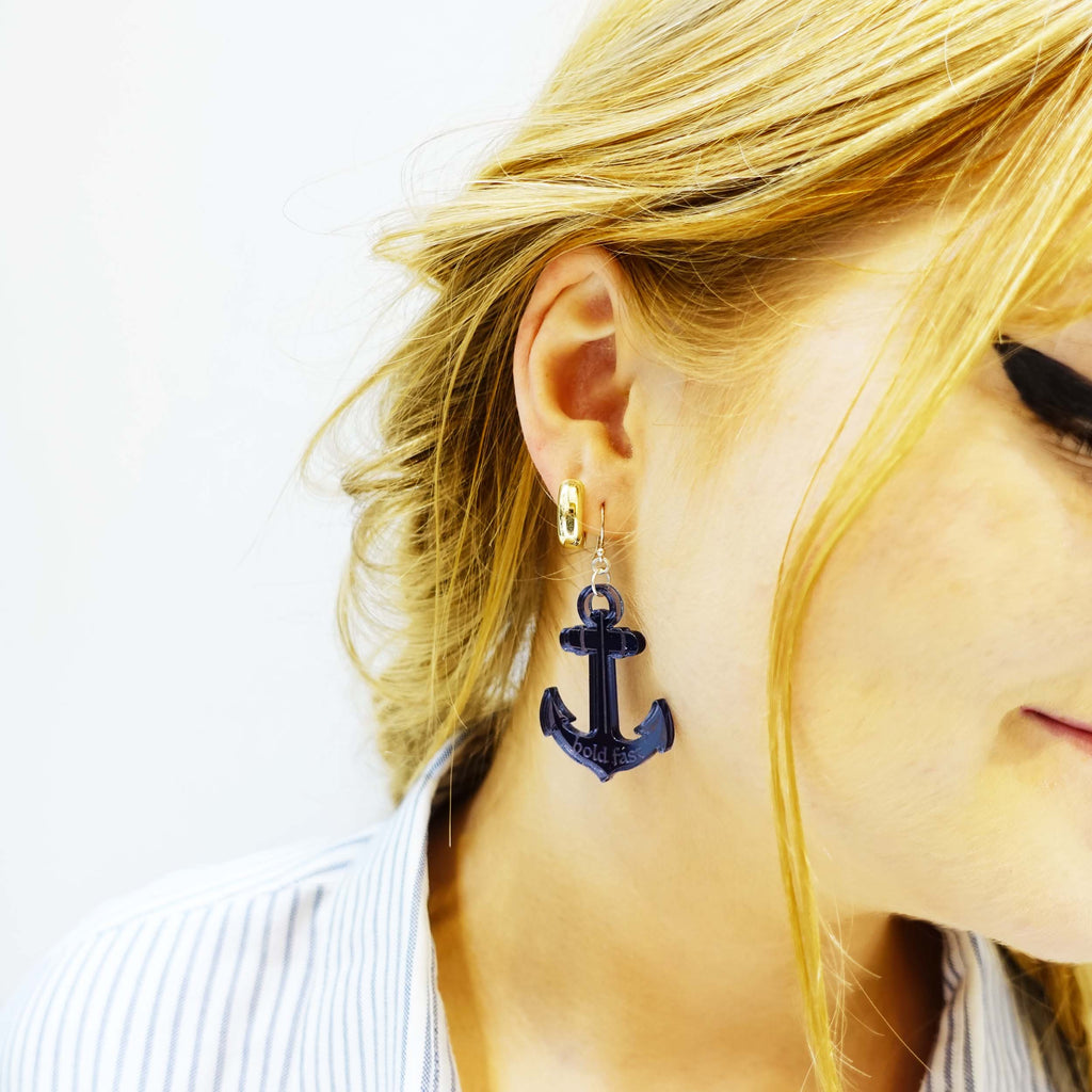Model wears slate anchor earrings, showing one etched with the words hold fast, with proceeds going to support the RNLI and Women for Refugee Women. 