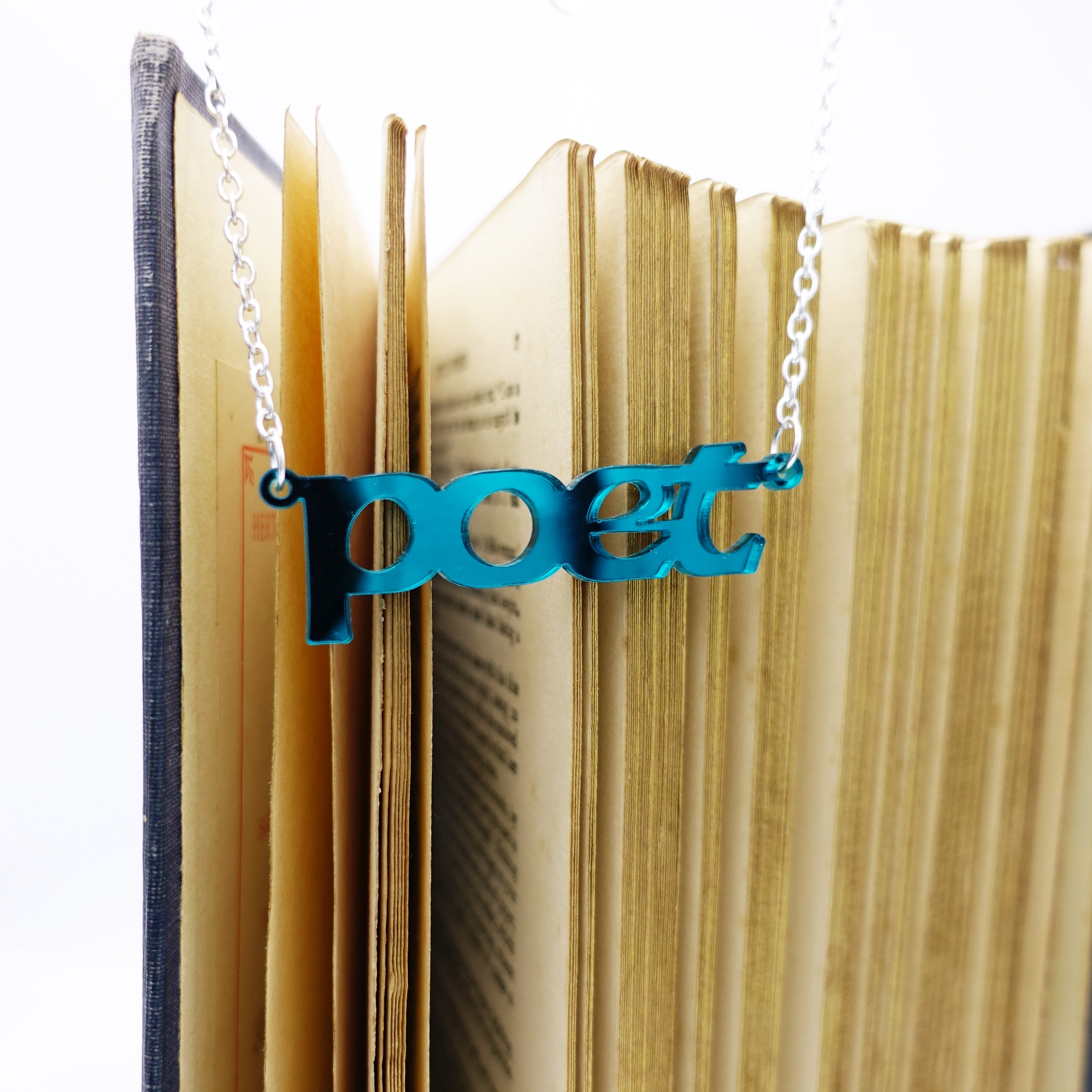 Poet necklace in teal mirror shown hanging with a book. 