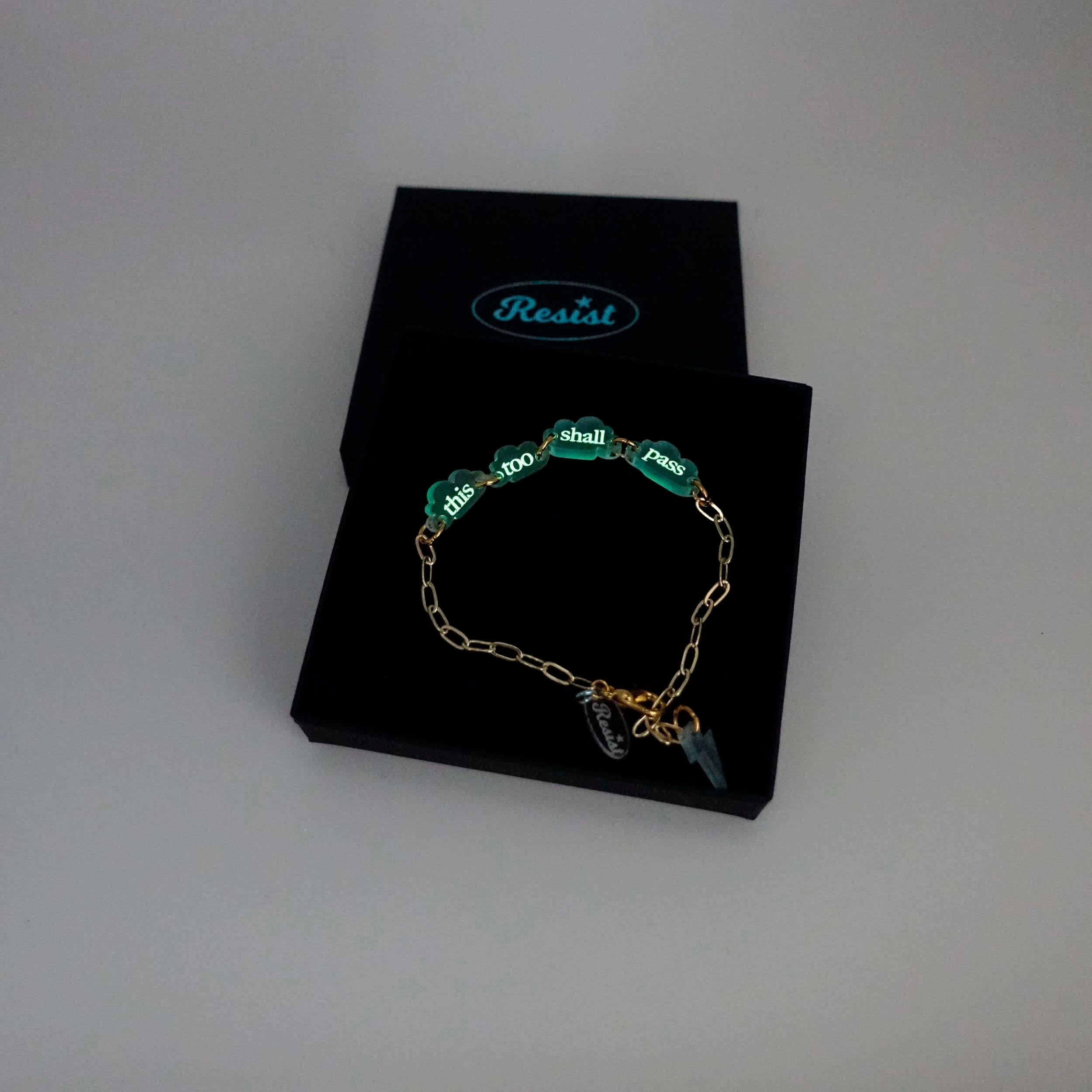 This too shall pass floating clouds gold-plated paperclip bracelet, etched with glow-in-the-dark pigment shown in a Wear and Resist gift box. 