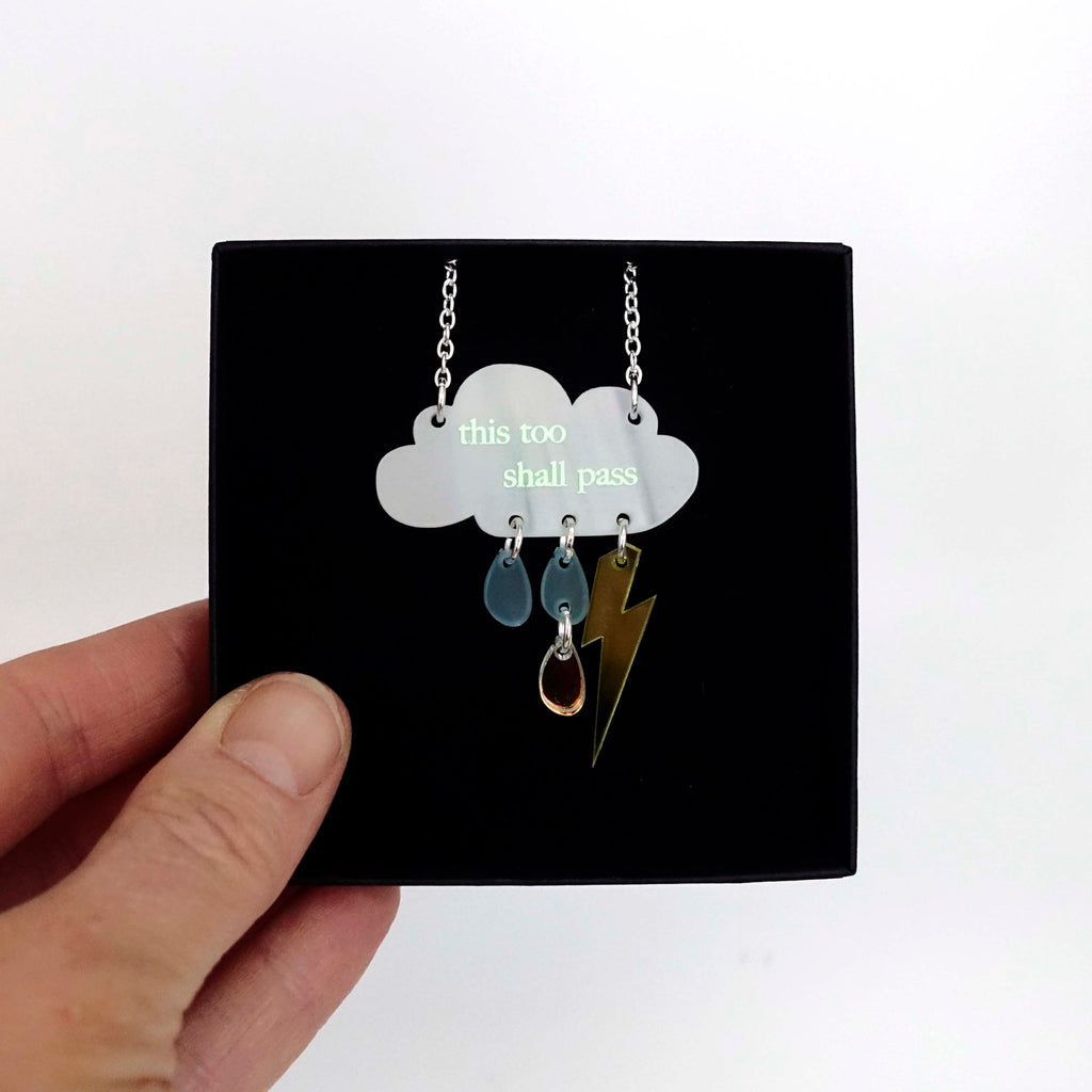 A 'This too shall pass' raincloud necklace etched with glow-in-the-dark pigment, shown held up in a Wear and Resist gift box. 